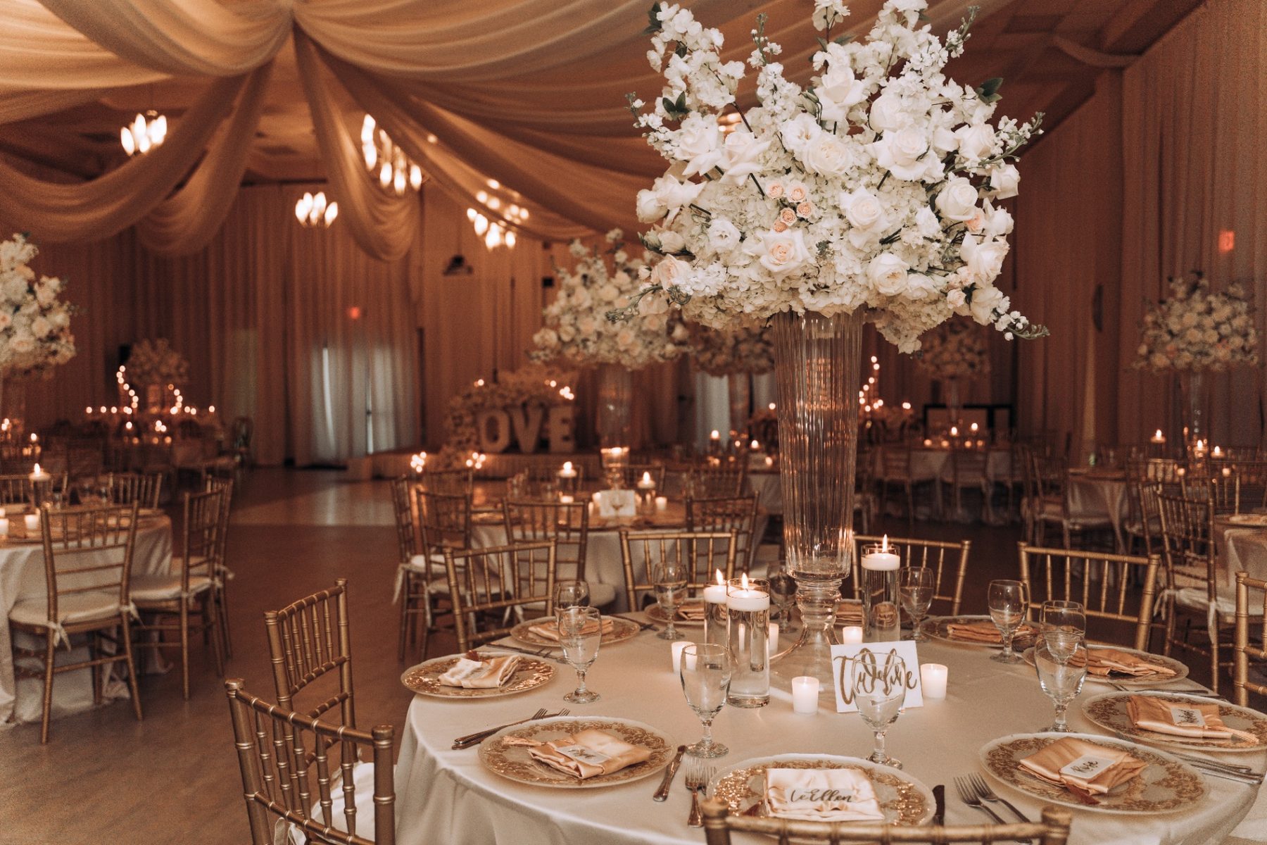 Gold ballroom wedding reception with tall rose centerpieces and glowing LOVE sign